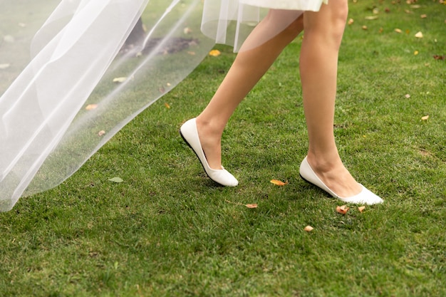 Bride in comfortable shoes and a long veil walks on the green grass on a wedding morning