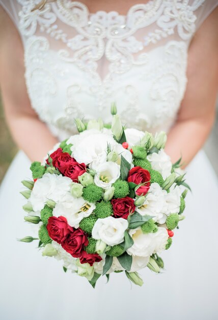 Bride in dress with lace corset holds white and red bouquet before her chest