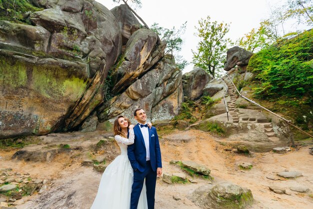 The bride embraces the groom from the back side and they smile picturesque rocks trees and stone stairs