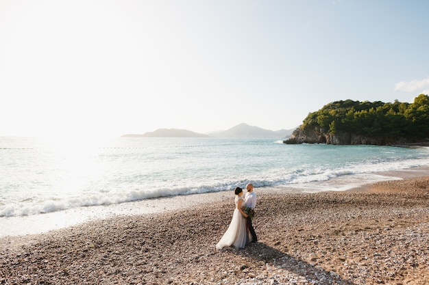 The bride and groom are hugging each other on the pebble beach by the sea