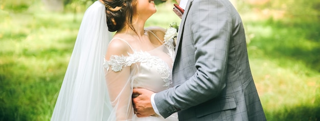 Bride and groom are hugging in the park. Happy couple walking together. Wedding day photo. Love story. Beautiful long sleeve dress. Lace veil. Stylish rustic bouquet.