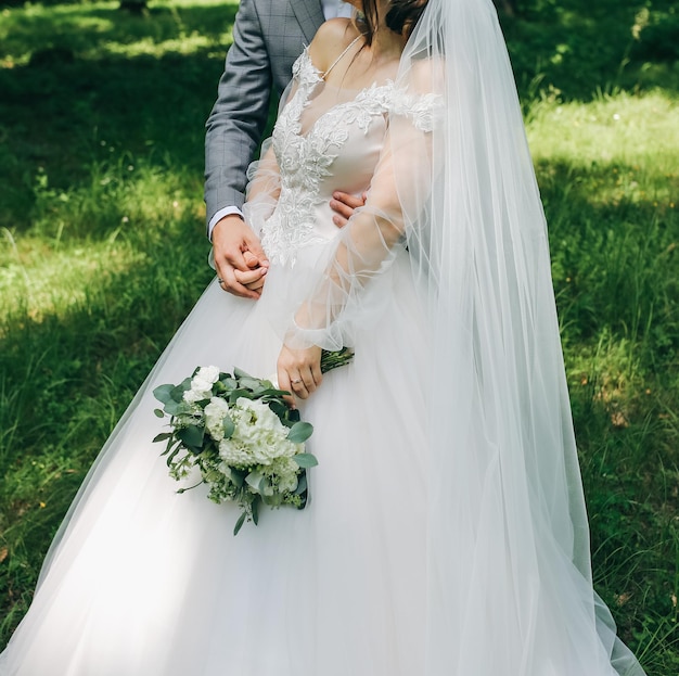 Bride and groom are hugging in the park Happy couple walking together Wedding day photo Love story Beautiful long sleeve dress Lace veil Stylish rustic bouquet