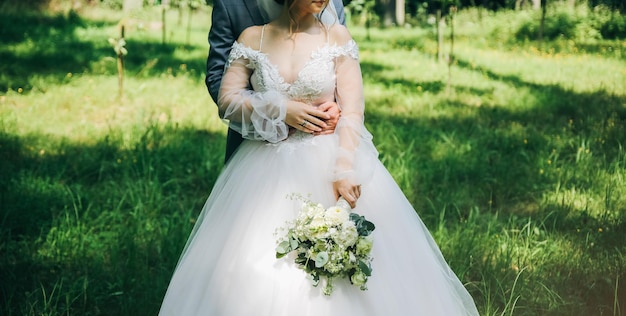 Bride and groom are hugging in the park Happy couple walking together Wedding day photo Love story Beautiful long sleeve dress Lace veil Stylish rustic bouquet