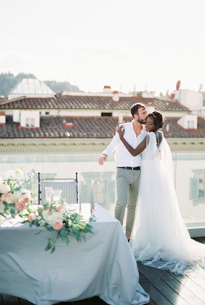 Bride and groom are hugging on the roof near the laid table with flowers