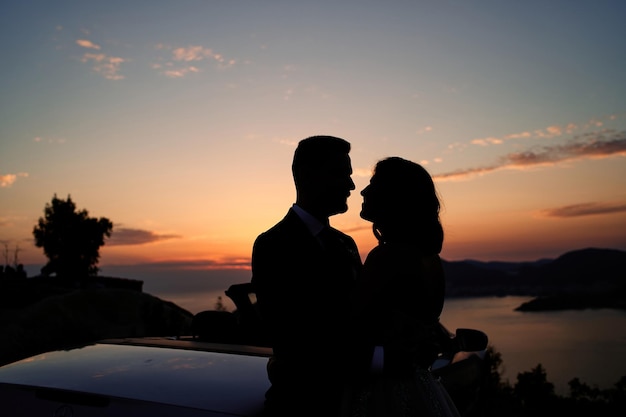 Bride and groom are hugging while looking at each other near a convertible in the mountains at