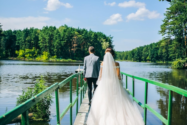 Bride and groom are walking on the bridge Wedding day photo near the lake and forest Love story Beautiful long sleeve dress Lace veil