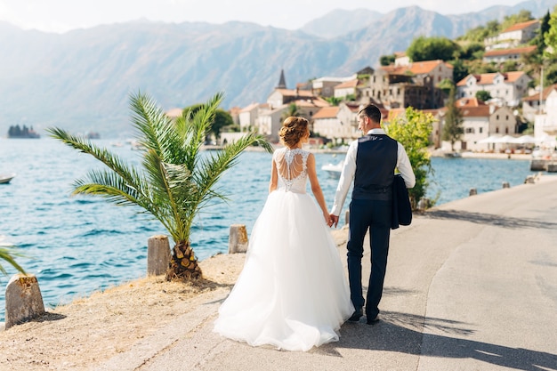 The bride and groom are walking hand in hand on the road by the sea near the old town of perast back