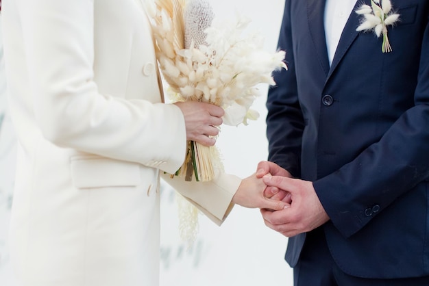 Bride and groom exchange wedding rings Bride in beige wedding suit with bouquet with dry flowers and groom in dark blue suit on their wedding day