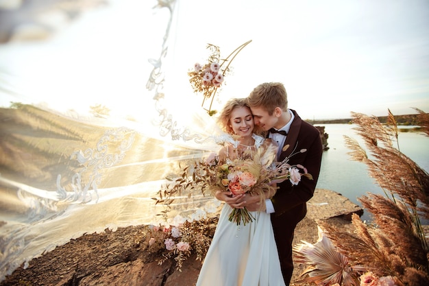 The bride and groom near the wedding decoration at a ceremony on a cliff of rock near the water at sunset. Veil flying from the wind