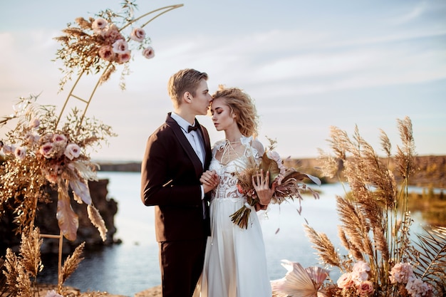 Bride and groom near the wedding decoration at a ceremony on a cliff of rock near the water at sunset