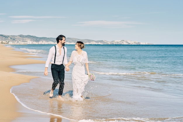 Bride and groom running along the beach holding hands and smiling happily celebrating their wedding.