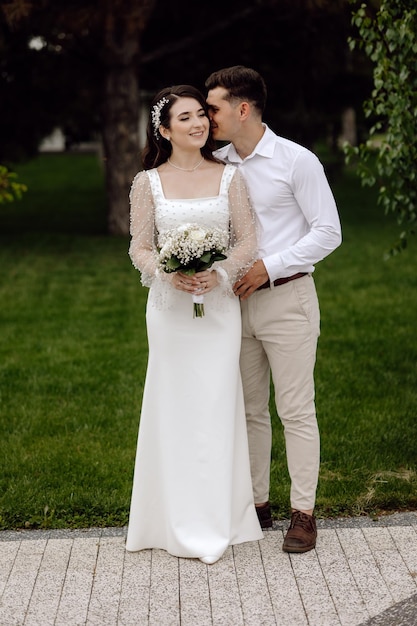 A bride and groom stand in front of a green lawn and the groom is wearing a white dress with a long sleeved neckline.