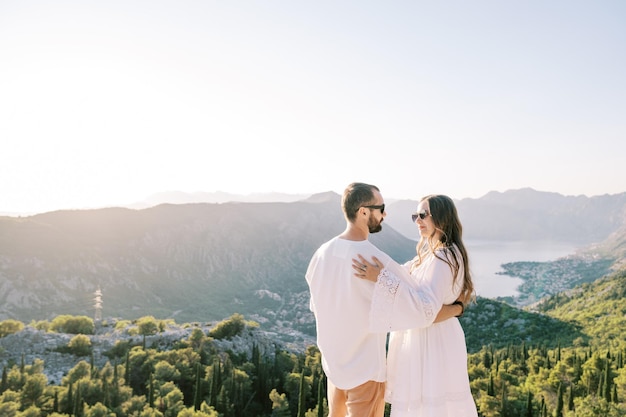 Bride and groom stand hugging and looking at each other on a mountain above the bay of kotor