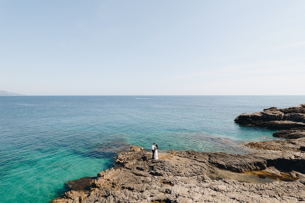 The bride and groom stand hugging on the rocky shore