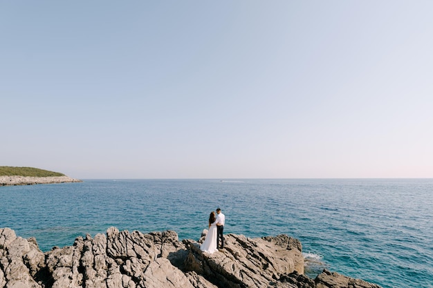 Bride and groom stand on the rocks and look at the sea back view