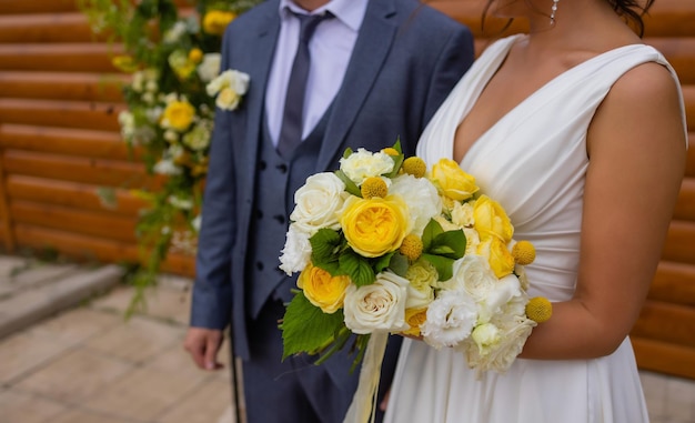 Bride and groom standing on green grass and holding a bouquet of white and yellow flowers with green