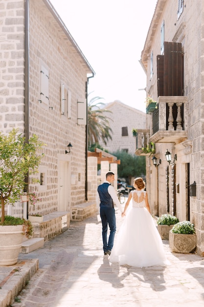 The bride and groom walk along a cozy narrow street of the old town of perast holding hands back