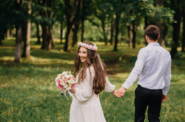 A bride and groom walk through a forest holding hands.