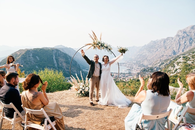 Bride and groom wave to the applauding guests standing at the wedding arch on the mountain above the