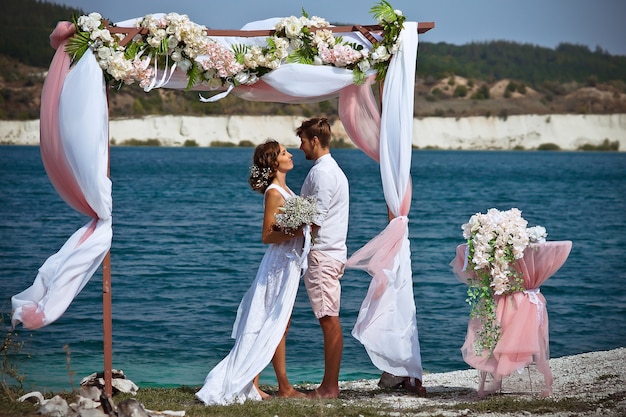 the bride and groom in white clothes with a bouquet of white flowers stand under an arch of flowers and fabric against the background of a blue lake and white sand