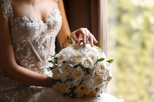 A bride holds a bouquet of roses in front of a window.