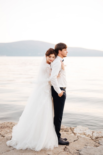 The bride hugs the groom, resting her head on his back on the pier near water