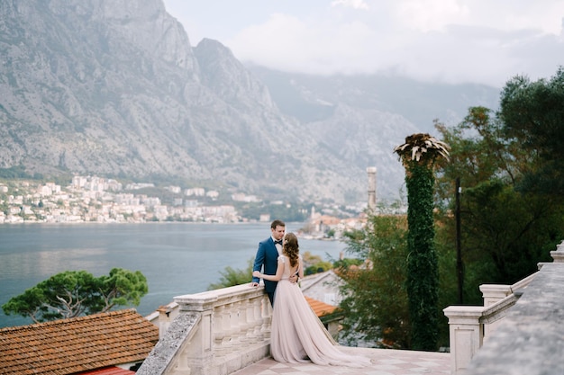 Bride hugs groom on the stone terrace against the backdrop of the bay