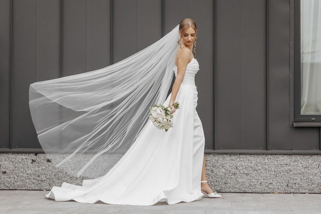 A bride is walking down a street with a white veil and a bouquet of flowers