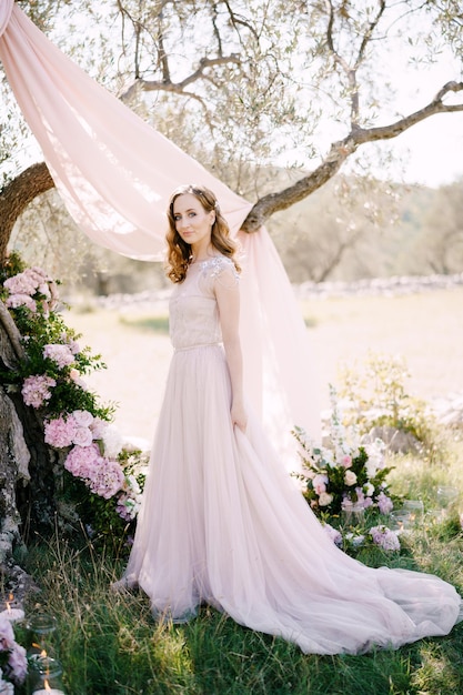 Bride in a long dress stands near a tree decorated with flowers