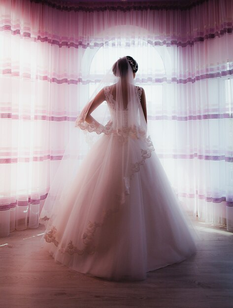 Bride in puffy tulle dress and wedding veil is standing near the window.