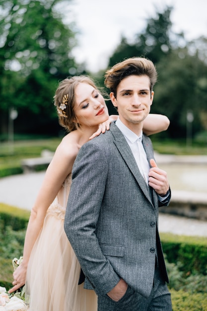 Bride put her hand on grooms shoulder hugging him from behind in front of the fountain