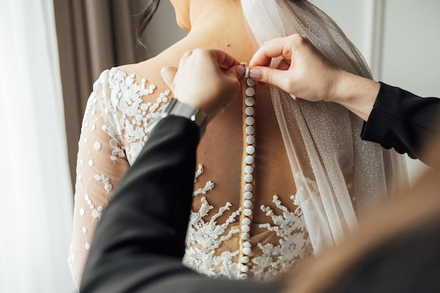 The bride puts on a white lace wedding dress mom and girlfriend of the bride help to tie the dress from the back