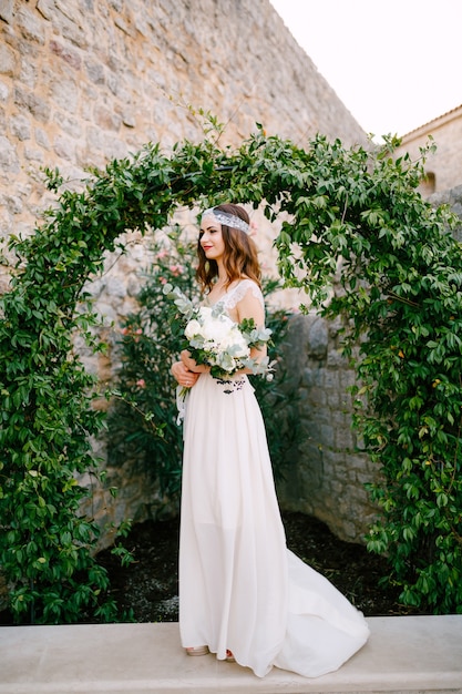 A bride stands at a graceful arch entwined with wild grapes in the old town of Budva and holds a bouquet in her hands