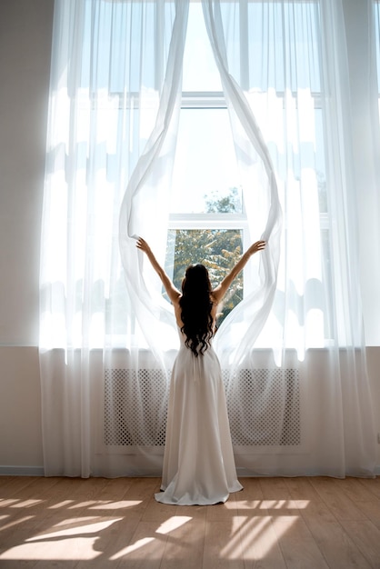 The bride stands with her back at the window opens the curtains in the hotel in a white wedding dress