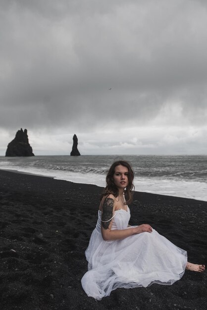 bride in white clothes stands on a black sand beach near the Atlantic Ocean in Iceland