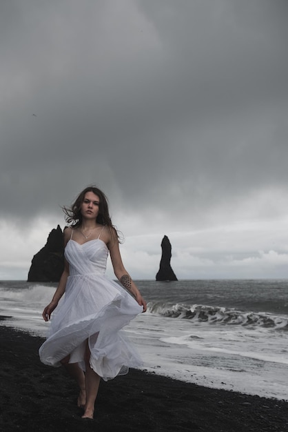 bride in white clothes stands on a black sand beach near the Atlantic Ocean in Iceland