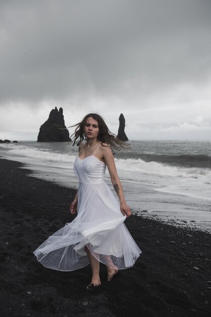 bride in white clothes stands on a black sand beach near the Atlantic Ocean in Iceland