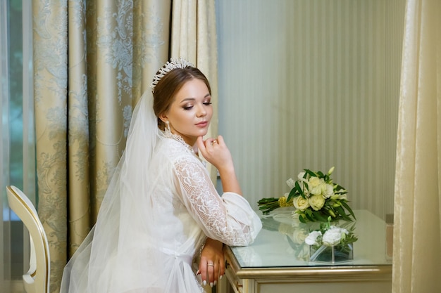 The bride in a white coat and veil sits at the makeup table