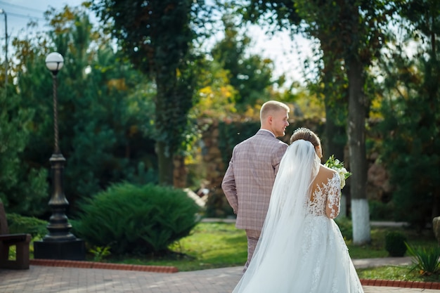 Bride in white dress and groom in costume cuddle and walk in the park