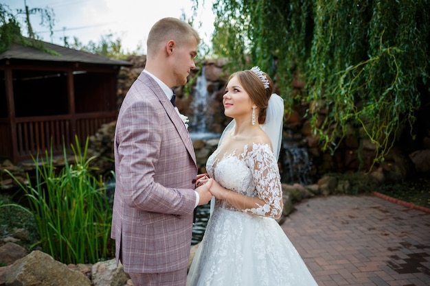 Bride in white dress and groom in costume cuddle and walk in the park