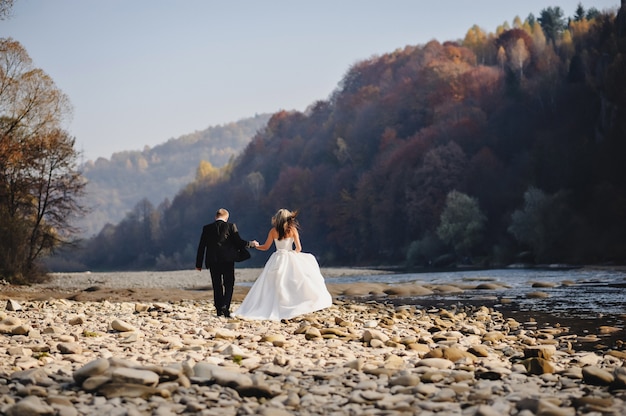 Bride in a white dress and groom go to the river rocks and holding hands