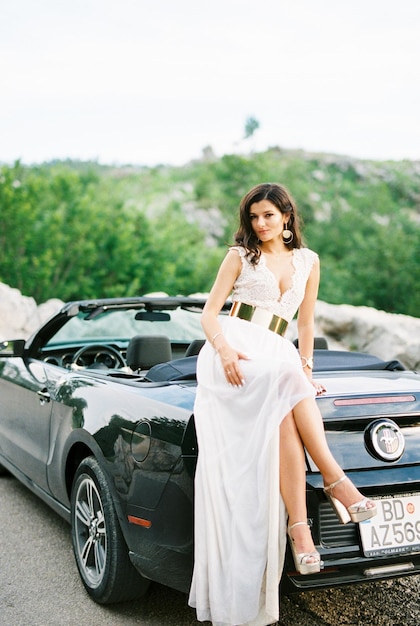 Bride in a white dress sits crosslegged on a sports convertible