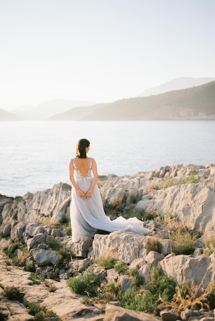 Bride in a white dress stands on a rocky seashore and looks at the mountains Back view