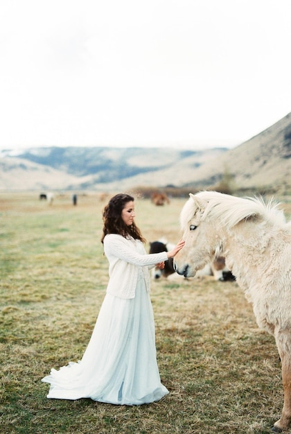 Bride in a white dress strokes a beige horse on the lawn Iceland