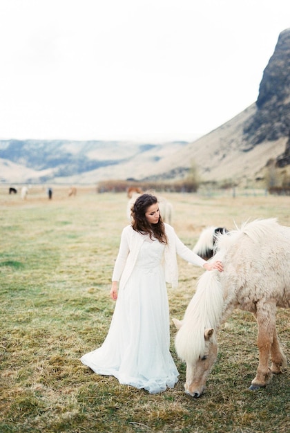 Bride in a white dress strokes the mane of a grazing horse