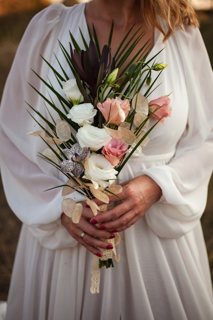 A bride in a white wedding dress holds a delicate bridal bouquet of roses and a lot of greenery. Stylish wedding bouquet on a sunset background