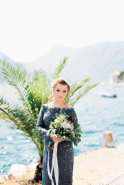 Bride with a bouquet of flowers stands on a pier by the sea against the backdrop of mountains