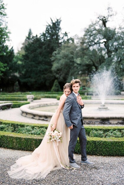 Bride with a bouquet hugs the groom from behind near the fountain