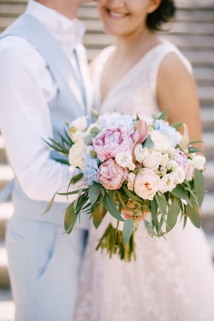 The bride with the bouquet of roses and peonies and groom stand hugging on the ancient stairs in