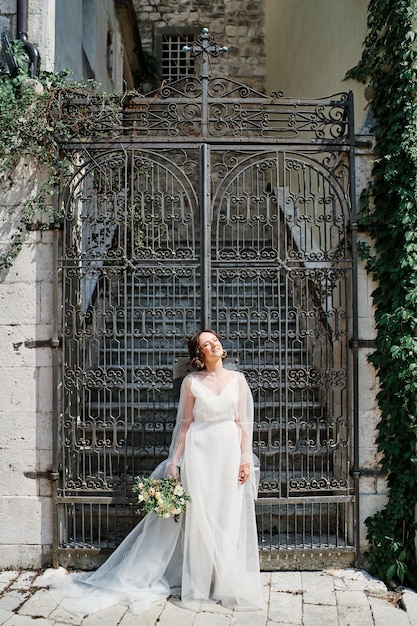 Bride with a bouquet stands leaning on the forged gate of an ancient building